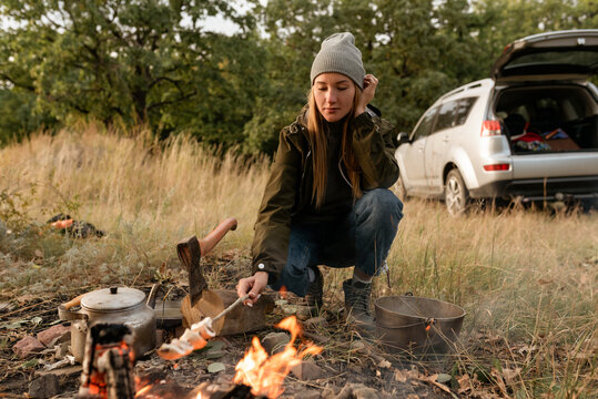 The blond girl sits near the firebond and fries mushrooms in the forest and the car stands behind her