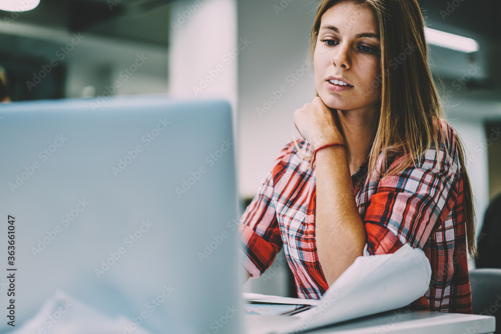 Poster pensive young woman dressed in casual wear watching tutorial online on website on modern laptop devi