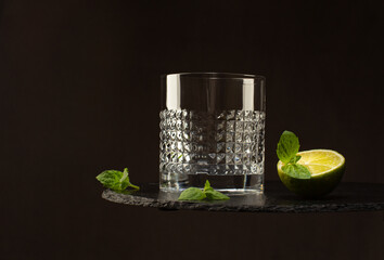 Empty crystal glass on black stone plate with fresh lime and mint leaves on dark background.Close-up.