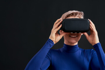Close up of excited woman with short hair in blue turtleneck wearing virtual reality headset, vr glasses isolated over dark background