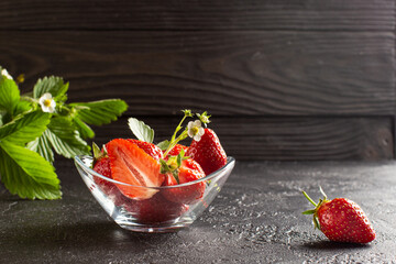 Fresh ripe strawberry in a glass bowl with strawberry flowers and leaves on dark background.