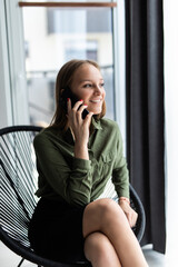 Fototapeta premium Portrait of a young professional girl sitting smiling at home using a smartphone mobile phone to have a phone conversation next to a large window.