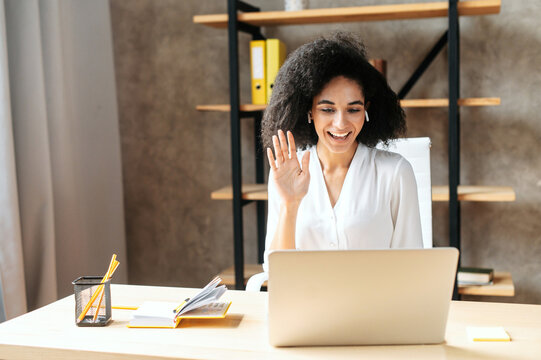 Cheerful biracial young woman uses laptop for video call. A glad girl sits in modern office and waving hello into webcam. Front view. Video meeting concept
