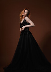 Beautiful elegant young model with bright redhead hairstyle posing in fashion chic black wedding dress with long skirt on studio grey background. Portrait