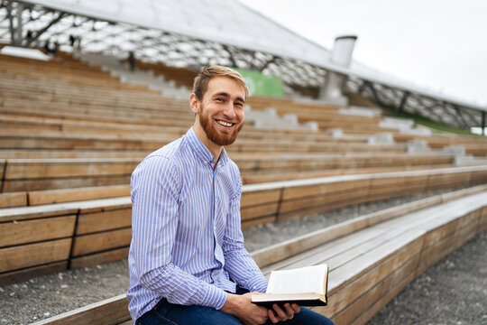 Cheerful male reading book on wooden bench at stadium