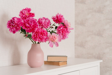 big bouquet of  peonies in a pink vase in a bright room are on a white chest of drawers, books are nearby. cozy house, fresh flowers at home, home greenhouse, minimal design, loft house