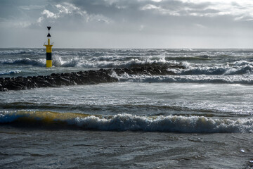 Dramatic lighting seascape with dark cloudy sky on a windy day churning waves, and a yellow signal pillar on a rock breakwater