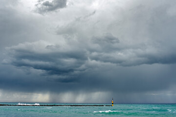 seascape, with dark and cloudy sky, raining, and the the sea in green tones, and a yellow signal pillar on a rock breakwater