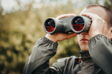 A young hunter in the forest looks into the binoculars. He is looking for wild animals.