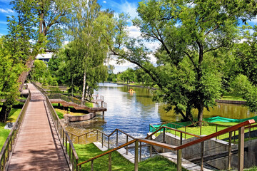 Fototapeta premium city park landscape at daytime with wooden pathway along pond embankment against blue sky with clouds background. Summer in Moscow city Russia