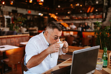 Fototapeta na wymiar A middle-aged caucasian casual businessman with glasses in a white shirt is sitting in a modern cafe, working on a laptop, drinking espresso coffee