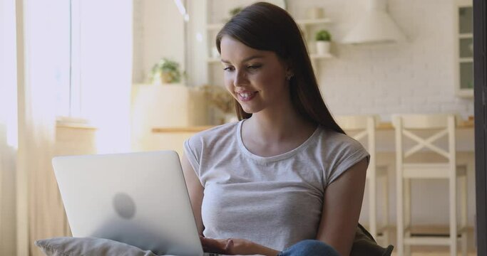 Smiling young woman using laptop notebook sitting at home, happy girl freelancer student typing on computer browsing internet working or studying online surfing web looking at screen in kitchen