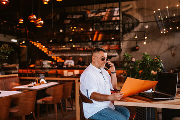 Fototapeta na wymiar A middle-aged caucasian businessman with glasses in a white shirt is sitting in a cafe, talking on the phone and reading business documents