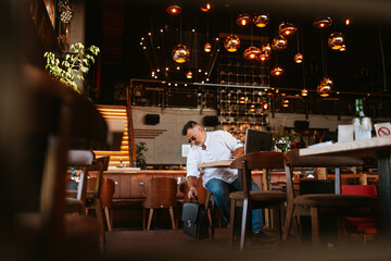 A middle-aged businessman with glasses in a white shirt sits in a modern cafe, works on a laptop and lifts a business bag from the floor