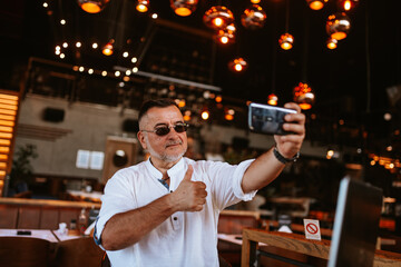 A middle-aged caucasian businessman with glasses in a white shirt sits in a cafe and takes a selfie while a laptop is on the table