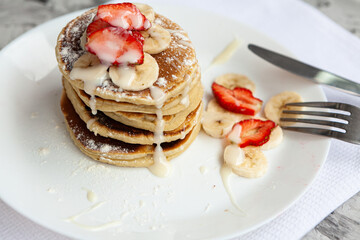 A stack of pancakes with fresh strawberries on a white plate on a light background. A white flower in a glass vase stands next to the Breakfast.
