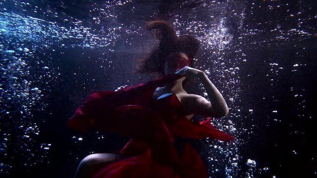 Close-up of a girl who is under water, she is wearing a long red dress that flutters on the water. There is a fabric floating around it and a lot of bubbles. She smiles and makes beautiful movements.
