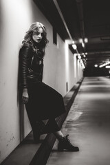 Obraz na płótnie Canvas Black and white photo of young female model in leather jacket. Woman leaning on white wall in background area of commercial building.