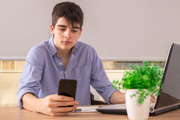 young male teenager or student at the desk with cellphone and computer