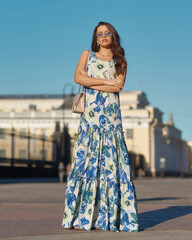 Elegant woman wearing white and blue colorful summer dress and sunglasses and walkig city street. Evening full length portrait. Fashionable female model posing outdoors