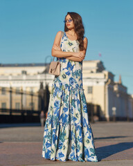 Naklejka premium Elegant woman wearing white and blue colorful summer dress and sunglasses and walkig city street. Evening full length portrait. Fashionable female model posing outdoors