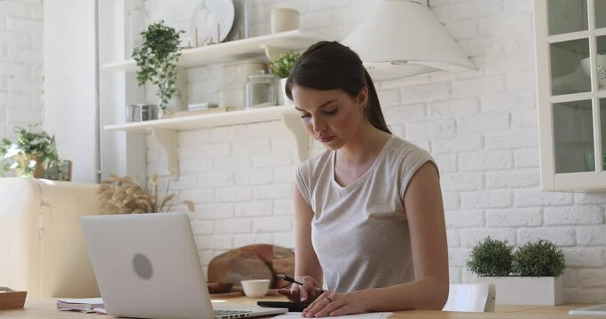Young smiling woman renter calculating paying bills using laptop computer calculator doing paperwork planning budget expenses managing money finances loan rent payments sit at kitchen table at home