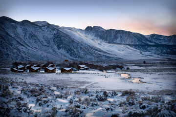 Snow covered mountains with small cabins nestled in the frozen valley next to a river reflecting the sunset