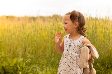 Little cute girl 1-3 with a plush hare in a light dress waving his hand in a field of spikelets of...