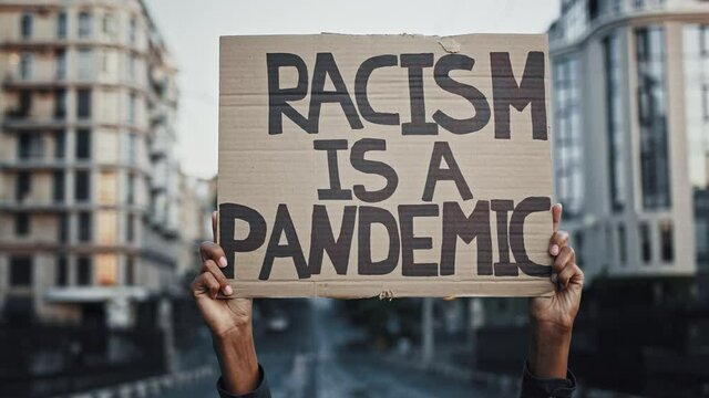Hands of unknown african-american person are raising up cardboard poster racism is a pandemic, posing against cityscape. Protests in USA. Close up