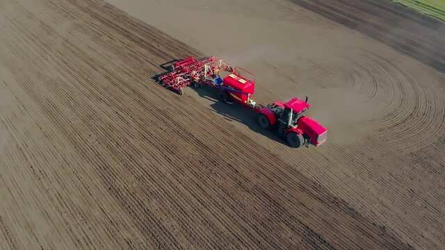 A red agricultural tractor with a seeder plows the land and planting seeds of grains in the fertilizer soil of an agricultural field. Spring work on farmland. Top view from the air, unmanned flight
