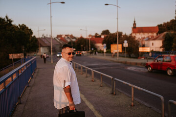 Fototapeta na wymiar A middle-aged caucasian businessman with glasses, in a white shirt and a black briefcase in his hand walks on the bridge