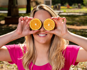 Happy and smiling young woman playing with an orange cut in half and in front of her eyes. Photo in...