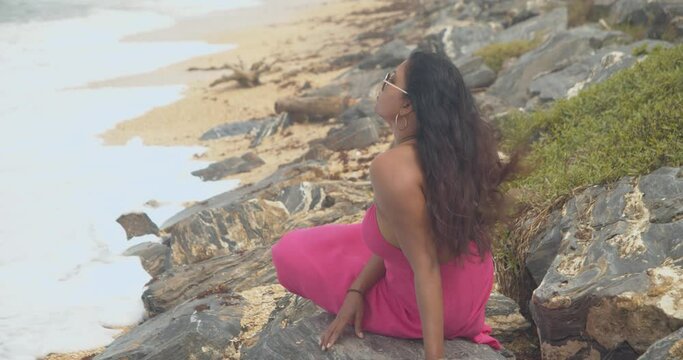 Amazing Caribbean model watching the waves of the ocean crash at her feet while siitting on the rocks