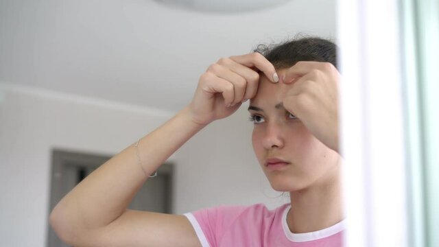 sad young woman pops zits on forehead with fingers looking at reflection in bathroom mirror standing at home close view