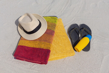 Top view of straw hat, yellow sunblock lotion bottle, flip flops and towels lie on white sand. Vacation concept in summer time.