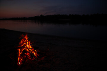 Bonfire on the river Bank at night. Red flames from burning branches and firewood near the reservoir in summer. Outdoor recreation by the fire.