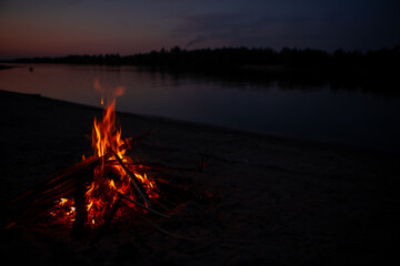 Bonfire on the river Bank at night. Red flames from burning branches and firewood near the reservoir in summer. Outdoor recreation by the fire.