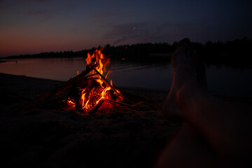 Men's feet on the river Bank near the fire at night. The light of fire illuminates the feet of a person. Red flames from burning branches and firewood near the reservoir in summer.