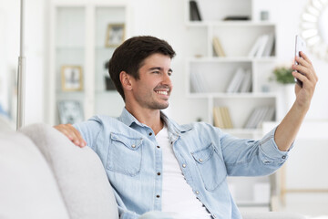 Handsome man sitting on a sofa at home communicates on his mobile phone making a video call or taking a selfie.