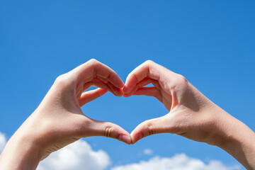 Close-up of hands making a heart symbol on a background of blue sky and clouds. Love, charity, valentines day concept - Powered by Adobe