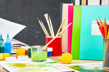 A textbook, acrylic and watercolor paints, brushes in a stand, colored school notebooks on a wooden...