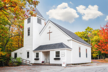 White country Baptist church surrounded by beautiful foliage colors in New England autumn. White clouds and blue sky background. - 363783836