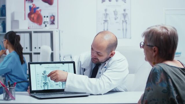 Young male doctor explaining how to avoiding bones problems to elderly retired old woman. Radiology and radiography in modern private hospital or clinic with medical staff walking in background, nurse