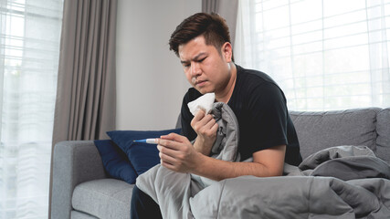 Sick Asian man with flu virus lying in sofa with high fever and measuring body temperature by thermometer and touching his forehead at home