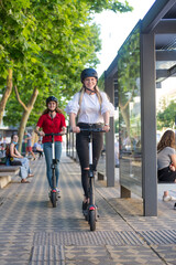 Fototapeta premium Trendy fashinable teenager girls riding public rental electric scooters in urban city environment. Eco-friendly modern public city transport.