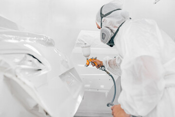 Worker in protective uniform spraying car body part in white paint.