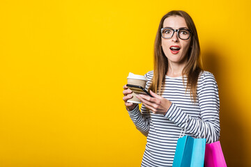 Surprised young woman with a phone, with shopping bags, with a on a yellow background.