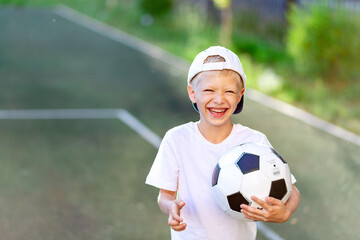 portrait of a laughing blond boy in a cap in a sports uniform with a soccer ball in his hands on the football field