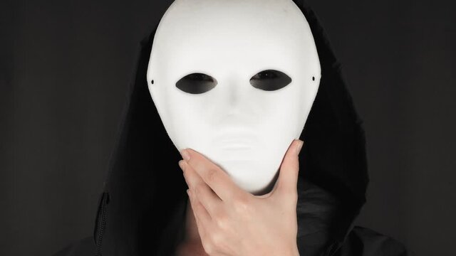 A woman with a make-up in a hood looks at the camera and takes off a white theater mask. Halloween face art