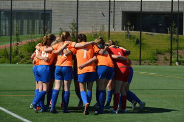 Female football players in orange shirts gathered in circle before the match to raise team spirit and discuss strategy. Selective focus.